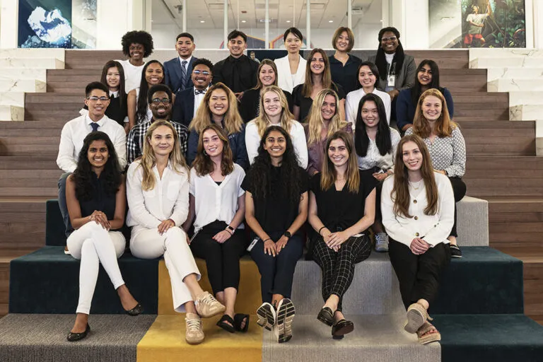 Image of a group of diverse young people dressed in business attire. They are inside, sitting on steps and smiling at the camera.