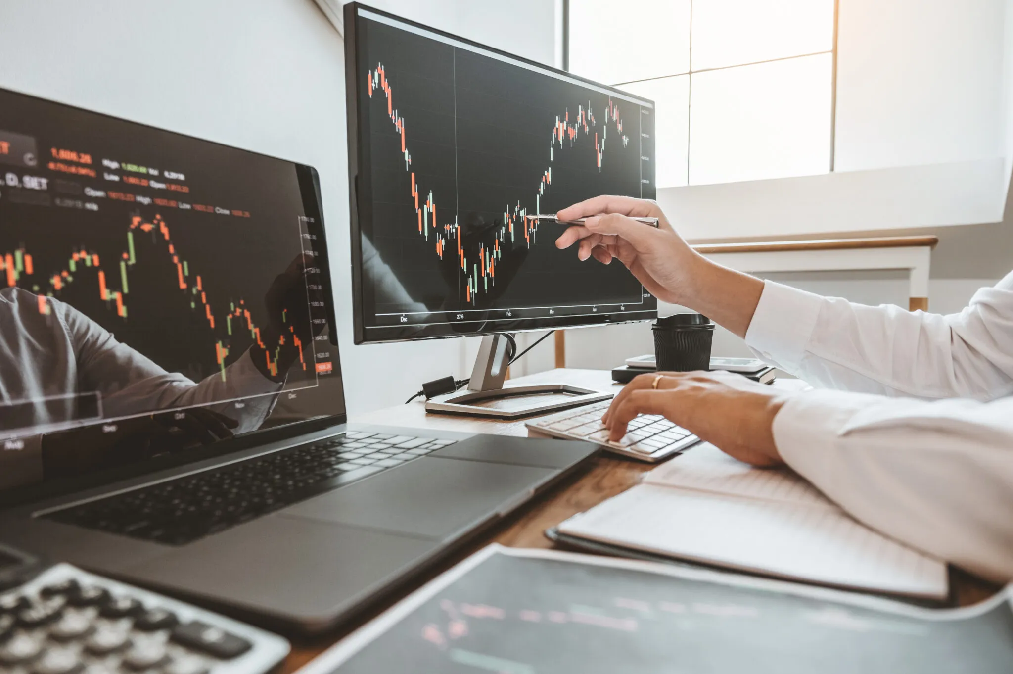 Two computer monitors showing graphs of growth over time. Someone’s hand is pointing at a portion of the graph while their other hand types information.