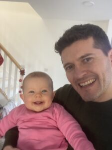 Picture of Cathal King. Cathal King presents as a White male. He has dark hair that it cut close. He holds his daughter Isla. Both are looking at the screen with big smiles.