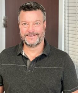 Troy Fornof is male presenting. He wears a casual, dark brown polo shirt, has dark hair that's graying at the temples, and he smiles broadly at the camera. 