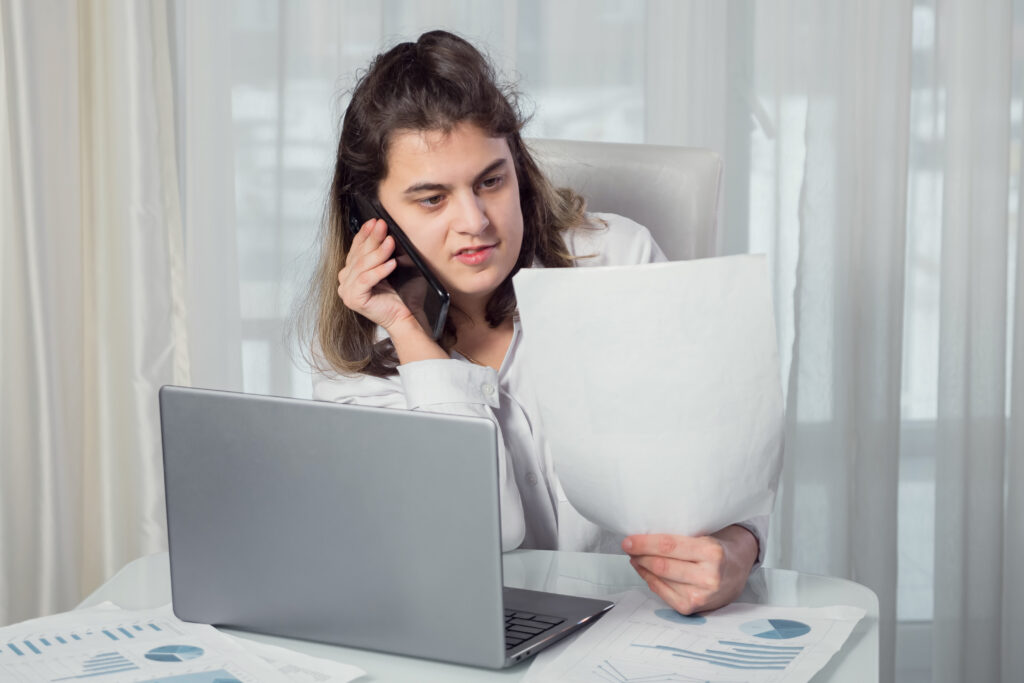 Female freelancer talks on a mobile phone explaining project paper near laptop at a table against a window at home.