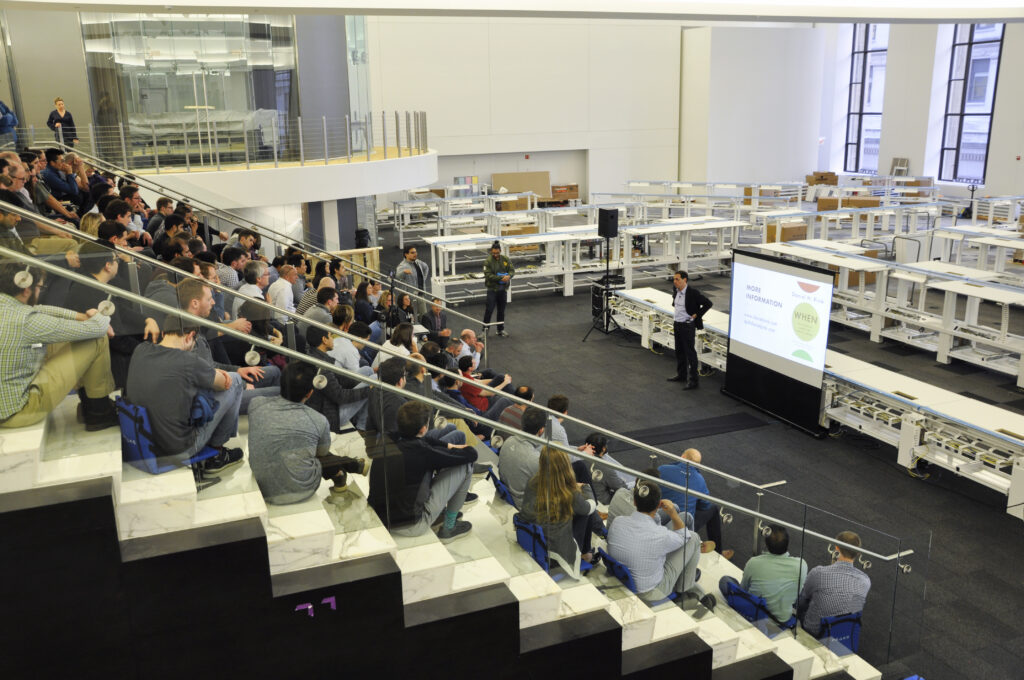 A speaker with a presentation stands in front of a large crowd of PEAK6 employees, all listening attentively.