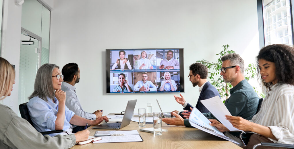 A diverse group of people on a business call, they are both in-person in a conference room, and another group is on a video screen calling in digitally.
